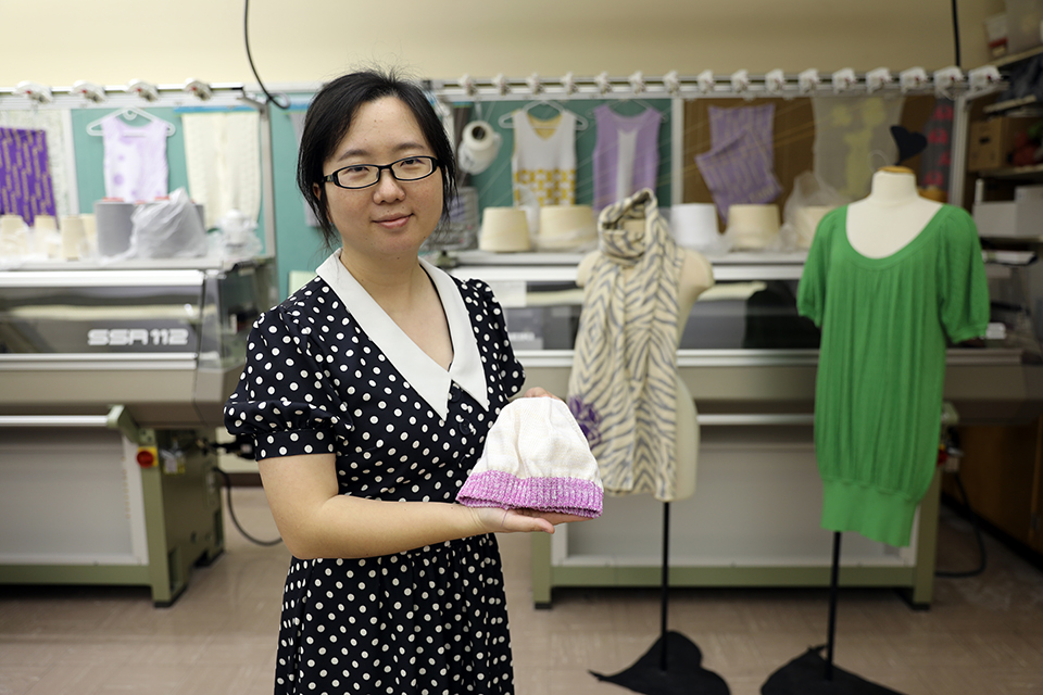 A woman holds a knitted cap in a room while in front of other garments.